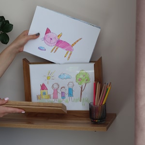 May include: A wooden picture frame with a white mat and a drawing of a family inside. The drawing is done in colored pencils and shows a family of four with a house and a sun in the background. There is a second drawing of a pink cat with yellow stripes on top of the frame.