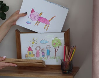 May include: A wooden picture frame with a white mat and a drawing of a family inside. The drawing is done in colored pencils and shows a family of four with a house and a sun in the background. There is a second drawing of a pink cat with yellow stripes on top of the frame.
