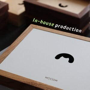 May include: A light wood frame with a white background and a black silhouette of a phone receiver. The text "WOODM" is printed in the bottom right corner of the frame. The text "In-house production" is written in green letters above the frame.