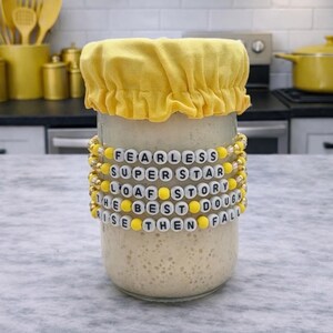 May include: A glass jar with a yellow fabric cover and beaded bracelets. The bracelets spell out "FEARLESS SUPERSTAR LOAF STORY THE BEST DOUGH RISE THEN FALL". The jar is filled with a light-colored mixture, likely dough.