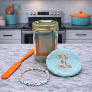 Pode incluir: Um frasco de vidro transparente com tampa dourada, cheio de uma subst&acirc;ncia de cor clara, est&aacute; sobre uma bancada ao lado de uma esp&aacute;tula laranja, uma pulseira de contas e um porta-copos turquesa com o texto "THE LOAF OF A - DOUGHGIRL".