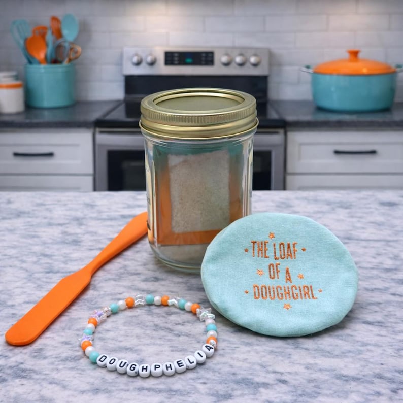 Pode incluir: Um frasco de vidro transparente com tampa dourada, cheio de uma subst&acirc;ncia de cor clara, est&aacute; sobre uma bancada ao lado de uma esp&aacute;tula laranja, uma pulseira de contas e um porta-copos turquesa com o texto "THE LOAF OF A - DOUGHGIRL".