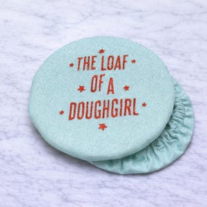 May include: A light blue, round fabric item with the text "THE LOAF OF A DOUGHGIRL" in red glitter lettering. The item has a matching gathered edge, and is likely a bread warmer or cover.