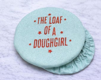 May include: A light blue, round fabric item with the text "THE LOAF OF A DOUGHGIRL" in red glitter lettering. The item has a matching gathered edge, and is likely a bread warmer or cover.