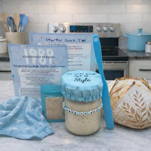 May include: A sourdough starter kit with instructions, a jar with a beaded bracelet that says "Bake it Off", a blue spatula, and a loaf of bread. The jar has a blue lid with the text "We Never Dough Out of Style".