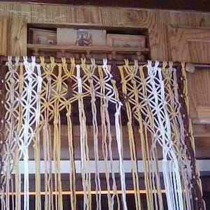 May include: A macrame curtain made with brown, white, and yellow yarn hangs from a wooden rod. The curtain is made with a knot pattern and has a fringe at the bottom.