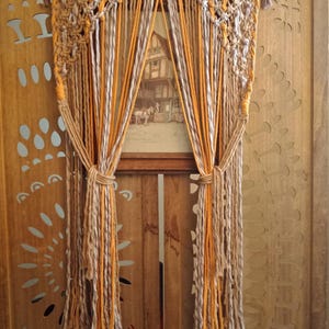 Könnte beinhalten: Ein Makramee-Vorhang in Beige-, Grau- und Orangetönen, der ein gerahmtes Bild einrahmt. Der Vorhang weist komplizierte Knotenmuster und lange, vertikale Stränge auf. Der Hintergrund ist eine Holztür mit dekorativen Ausschnitten.