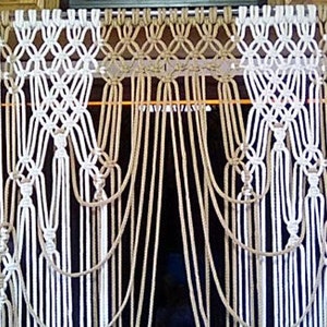 May include: A macrame wall hanging with a white and beige rope design. The hanging is made of multiple strands of rope that are knotted together to create a decorative pattern.