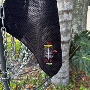 May include: Black microfiber towel with an embroidered disc golf basket and disc, with the letters "AR" below.