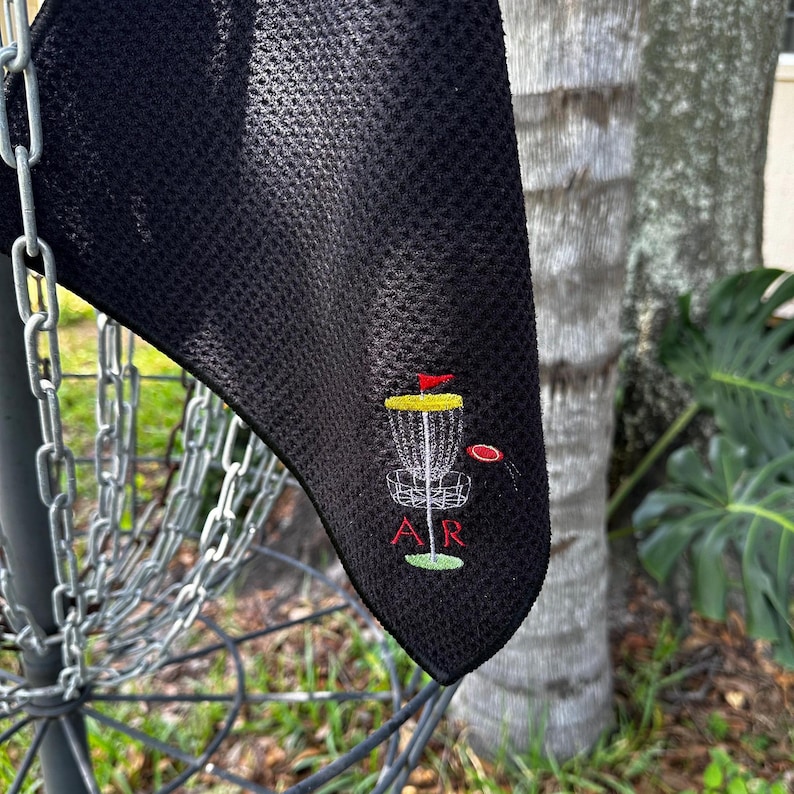 May include: Black microfiber towel with an embroidered disc golf basket and disc, with the letters "AR" below.
