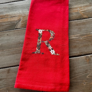 May include: Red kitchen towel with a white embroidered letter "R" surrounded by green leaves and white flowers.