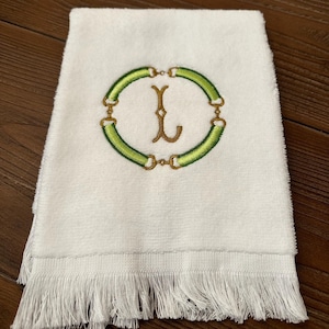 May include: White hand towel with a fringed edge. The towel features an embroidered design of a green oval with gold accents and a gold letter "L" in the center. The towel is folded.