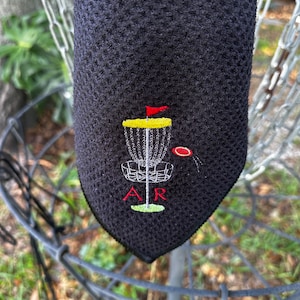 May include: Black microfiber towel with an embroidered disc golf basket, a red flag, and the letters "AR".