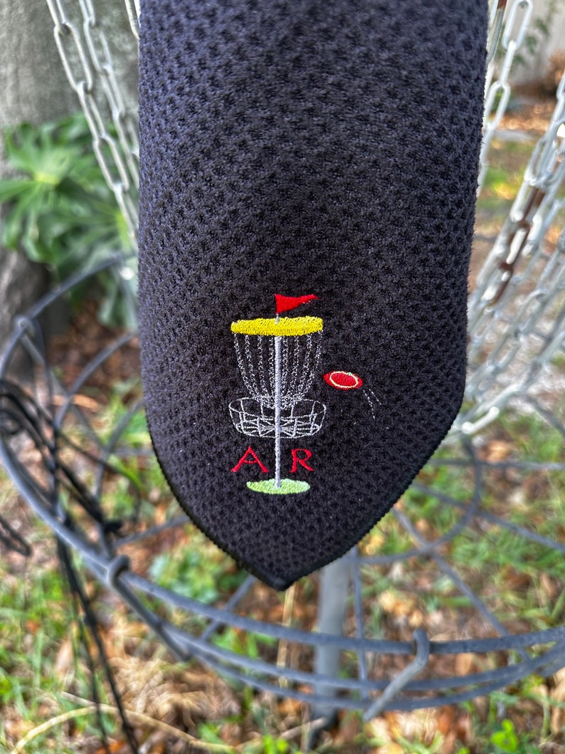 May include: Black microfiber towel with an embroidered disc golf basket, a red flag, and the letters "AR".