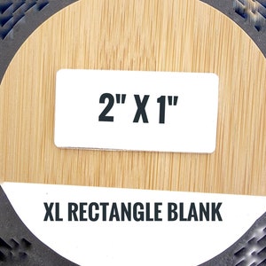 May include: A white rectangular label with the text "2" X 1" in black. Below the label, the text "XL RECTANGLE BLANK" is displayed in black. The label is on a light brown, wood-grain surface.