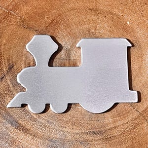 May include: A silver-colored train-shaped metal cutout. The cutout features a detailed design of a classic steam engine, including a smokestack, cab, and wheels. The cutout is placed on a wooden surface.