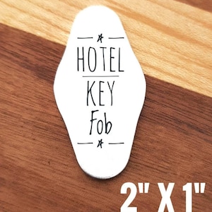 May include: White hotel key fob with black text that reads "HOTEL KEY Fob". The key fob is 2 inches by 1 inch. The key fob is on a wooden surface.