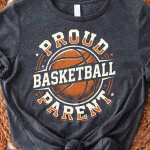 Puede incluir: Camiseta gris oscuro con un gráfico de baloncesto. El diseño presenta las palabras "PROUD BASKETBALL PARENT" en naranja y blanco, rodeando una pelota de baloncesto naranja. La camiseta es de material suave.