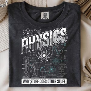 May include: Dark gray t-shirt with the word "PHYSICS" in large white and gray letters. The shirt features scientific diagrams, formulas, and the phrase "WHY STUFF DOES OTHER STUFF" in white text. The shirt is made of a soft material.
