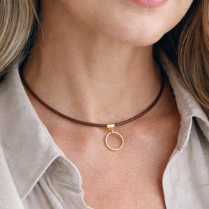 Puede incluir: Un collar gargantilla de cuero marrón con un colgante circular de color dorado. El collar se lleva sobre una camisa abotonada de color claro. El colgante es de diseño sencillo y elegante.