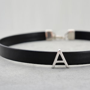 Może przedstawiać: Czarny skórzany naszyjnik typu choker ze srebrną zawieszką w kształcie litery "A". Naszyjnik ma prosty design z zapięciem. Litera "A" jest wyśrodkowana z przodu naszyjnika.