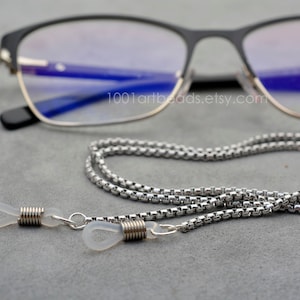 May include: A silver-coloured glasses chain with a box chain design. The chain has clear rubber loops to attach to the glasses. The glasses have a black frame with silver accents. The background is a solid grey colour.