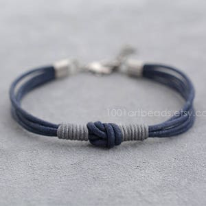 May include: A navy blue and gray braided cord bracelet with a knotted closure. The bracelet is made of multiple strands of cord and has a silver clasp.