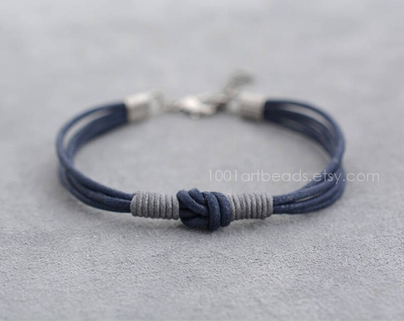 May include: A navy blue and gray braided cord bracelet with a knotted closure. The bracelet is made of multiple strands of cord and has a silver clasp.