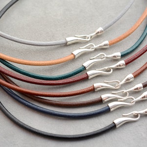 May include: A collection of leather necklace cords in various colors, including gray, tan, green, burgundy, brown, navy, and black. Each cord is fitted with a silver-colored clasp. The cords are arranged in a circular pattern, showcasing their different hues.