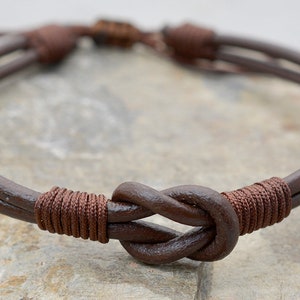 Könnte beinhalten: Ein braunes Lederarmband mit einem Knotendesign. Das Armband besteht aus zwei Ledersträngen, die mit einem Knoten zusammengebunden sind.