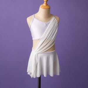 May include: A white two-piece dance outfit featuring a sleeveless crop top and a matching skirt. The set is accented with a sheer white fabric overlay that drapes diagonally across the torso, connecting the top and the skirt with thin straps.