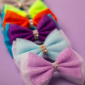 May include: A selection of vibrant hair bows in purple, blue, orange, and green, each with a sparkling rhinestone centre. The bows are arranged diagonally against a purple backdrop. The website address "cbcsportswear.com" is visible.