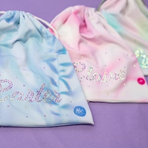Könnte beinhalten: Drei Beutel mit Kordelzug und Batikmustern in Blau, Pink und Grün. Jeder Beutel zeigt einen mit klaren Strasssteinen geschriebenen Namen, darunter Carter und Claire, sowie verstreute Strasssteine und ein kleines rundes Logo-Etikett.