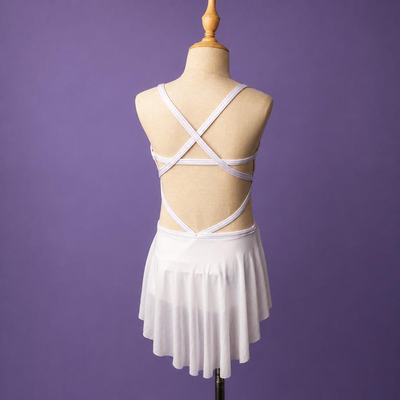 May include: A white leotard with a skirt, featuring a crisscross strap design on the back. The garment has a fitted bodice and a sheer, flowing skirt attached at the waist, displayed on a mannequin against a solid purple background.
