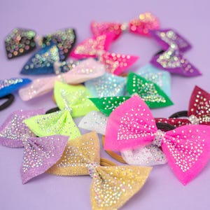 May include: A selection of vibrant hair bows, adorned with glitter and rhinestones. The bows are in shades of pink, blue, green, purple, and gold, set against a purple backdrop. Each bow is attached to a black elastic band.