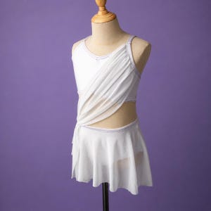 May include: A white dance leotard featuring a sleeveless bodice with a draped, semi-sheer overlay across the chest and a matching attached skirt. The garment includes thin straps and subtle silver trim along the neckline and waistline.