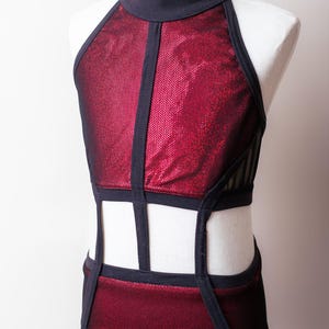May include: A sleeveless leotard featuring a high mock neck, constructed with metallic red textured fabric panels and solid black trim. The design includes cut-out sections at the waist and sides, connected by vertical black fabric bands.