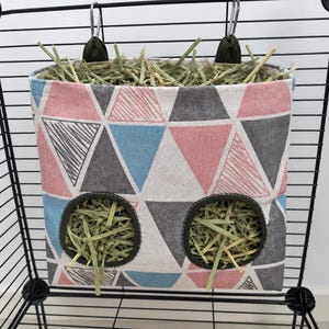 May include: A fabric hay feeder bag featuring a geometric pattern of triangles in shades of pink, blue, gray, and white. The bag has two circular openings with dark trim and is suspended by two metal clips from a black wire cage structure.