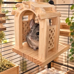 May include: A light-colored wooden chinchilla house with an arched roof and lattice side panels. A grey chinchilla is positioned inside the structure. The house is mounted on a black wire cage wall, featuring a flat wooden base platform.