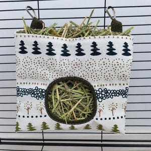 May include: A white fabric hay feeder bag with a repeating pattern of dark green trees, brown circles, and floral motifs. The bag features a circular opening in the center and is attached to a black wire cage with two olive green fabric straps.