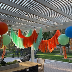 May include: Outdoor party decorations featuring orange, green, and blue balloons, and a fringed garland. A wooden table with black wicker chairs is in the foreground. A white pergola with string lights hangs above the decorations.