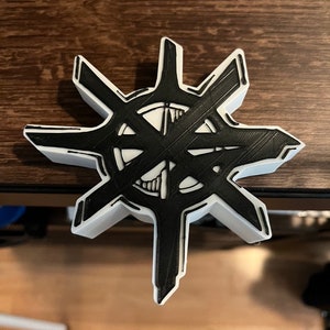 Puede incluir: Un objeto con forma de estrella impreso en 3D, con una combinación de colores blanco y negro. El diseño presenta un patrón geométrico complejo en negro, sobre un fondo blanco. El objeto está sobre una superficie de madera.