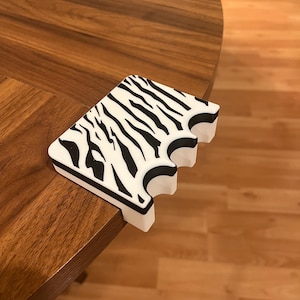 Puede incluir: Un protector de esquina blanco y negro con estampado de cebra descansa sobre una mesa de madera. El protector tiene una forma única con recortes curvos, diseñado para encajar en una esquina de la mesa. Las rayas negras contrastan con el fondo blanco.