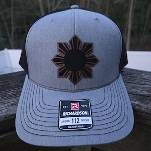 May include: Gray trucker hat with a black mesh back and a brown and black sunburst design on the front. The hat has a curved brim and an adjustable strap. The Richardson brand label is visible.