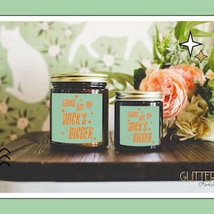 May include: Two glass jar candles with gold lids. Each candle has a teal label with the text "Turns Out My Wick's Bigger" in orange. The candles are on a dark wooden surface, with flowers in the background.