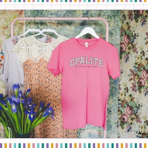 May include: A bright pink short-sleeved t-shirt with the word OPALITE printed in a white varsity-style font across the chest, hanging on a white hanger alongside a floral dress and a grey garment on a pink metal clothing rack.