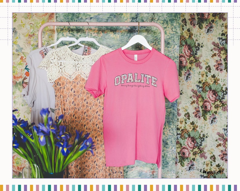 May include: A bright pink short-sleeved t-shirt with the word OPALITE printed in a white varsity-style font across the chest, hanging on a white hanger alongside a floral dress and a grey garment on a pink metal clothing rack.