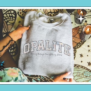 May include: A light gray Opalite sweatshirt with the text "OPALITE" in a block letter design. Below the word "OPALITE" is the text "dancing through the lightning strikes". The sweatshirt is held by a person.