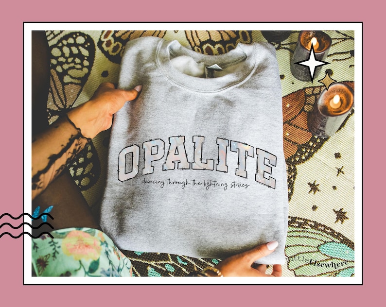 May include: A light grey crewneck sweatshirt featuring the word OPALITE in large, block-style letters with a barbed wire outline. Below the main text, the phrase dancing through the lightning strikes is printed in a smaller, cursive font.