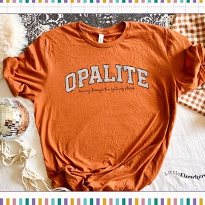 May include: A burnt orange short-sleeve t-shirt featuring the word OPALITE in large, distressed white block letters with a black outline, and the cursive text dancing through the lightning strikes printed underneath the main graphic.
